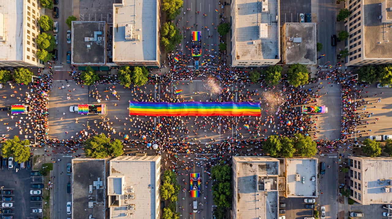 Aerial view of Houston Pride 2026 parade crowd lining downtown streets around a large rainbow flag carried by marchers.