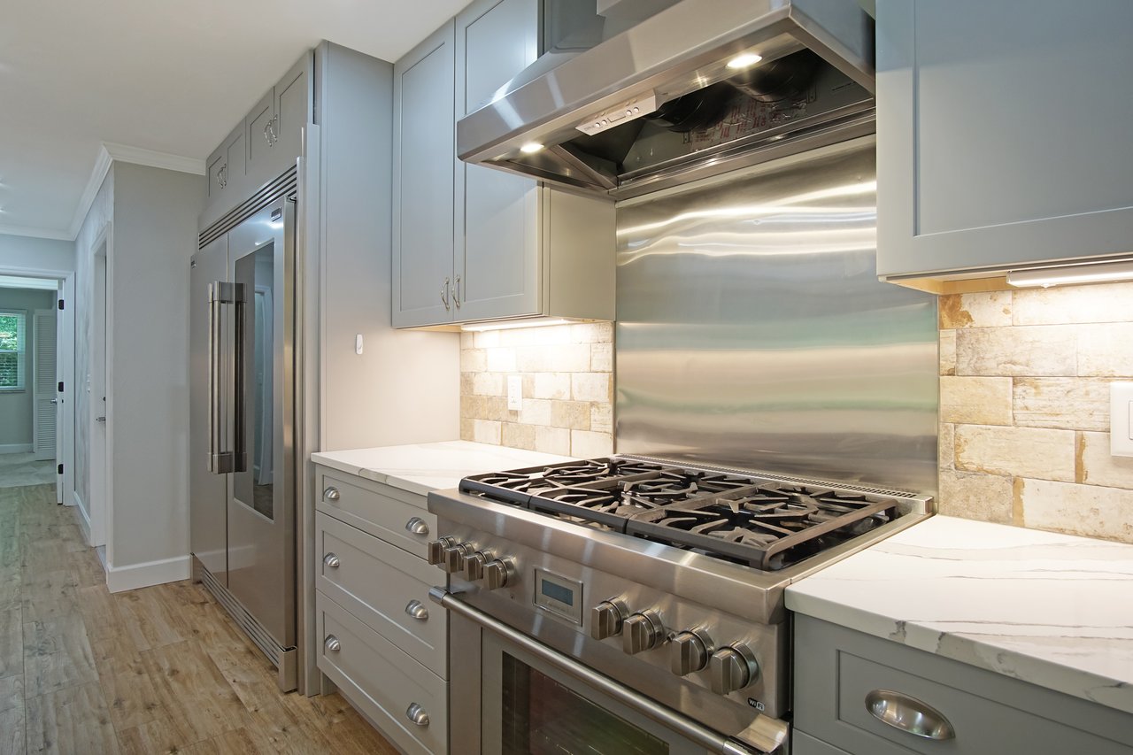Modern kitchen with stainless steel gas stove, light gray cabinets, and marble countertops. Light wood flooring leads to a hallway. Warm, inviting tone.
