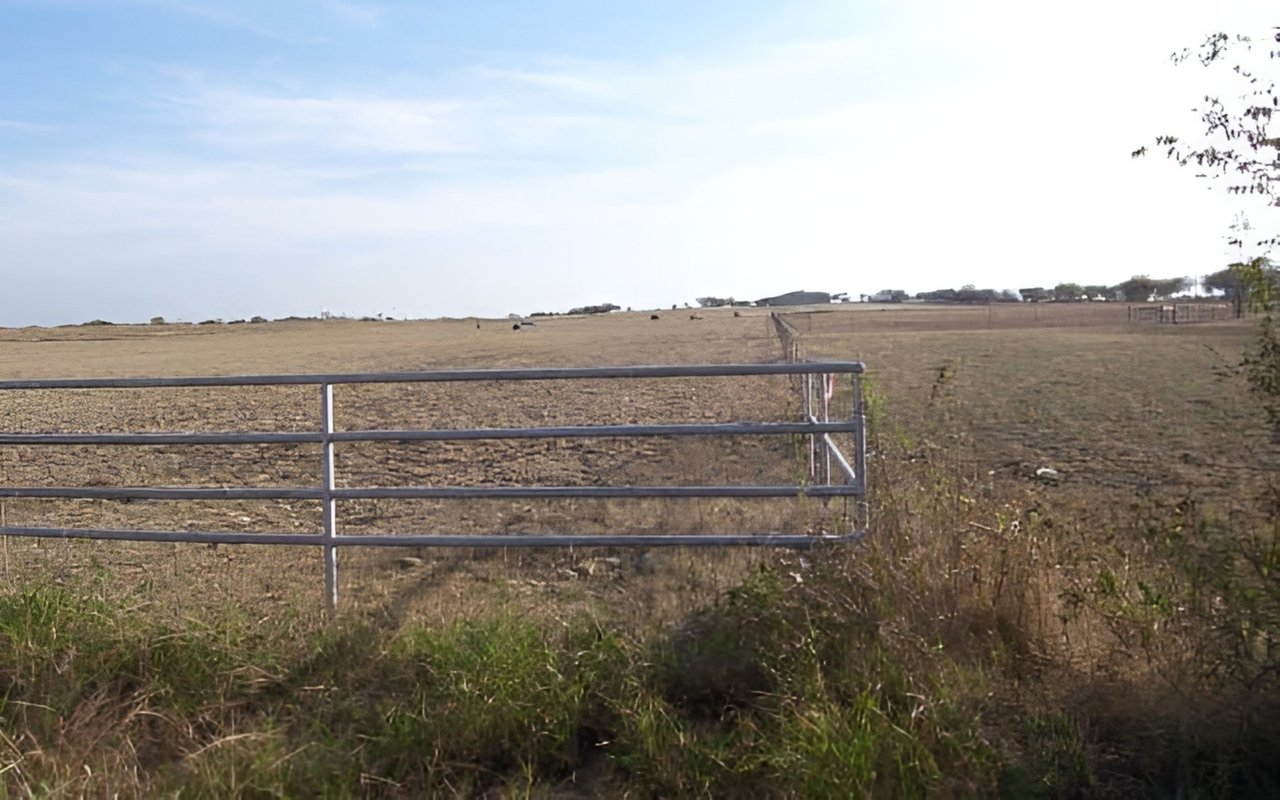 Open land behind a metal gate, with dry fields and distant buildings.