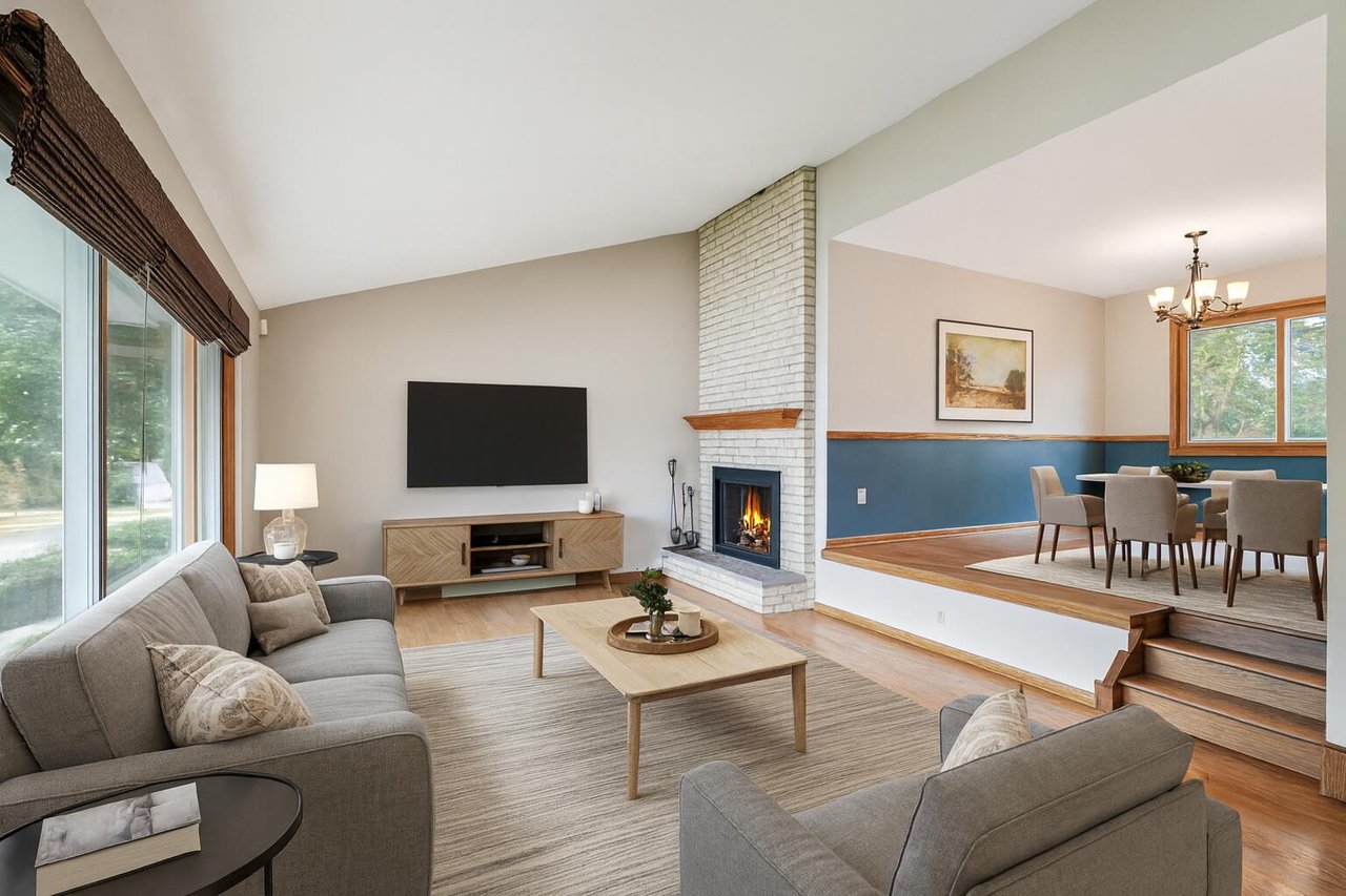 Bright modern living room with a fireplace, large windows, neutral décor, and an open elevated dining area.