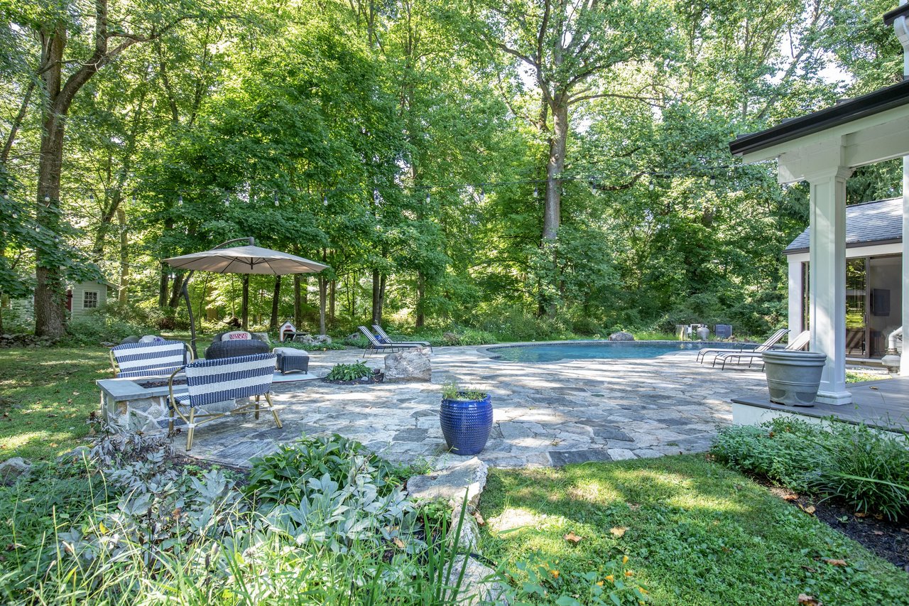 Backyard pool and stone patio with lounge seating beneath mature trees.