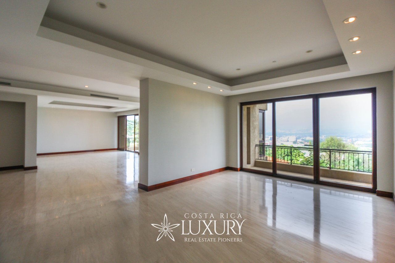 Vacant apartment living area with polished floors, tray ceiling, and balcony overlooking greenery and the city.