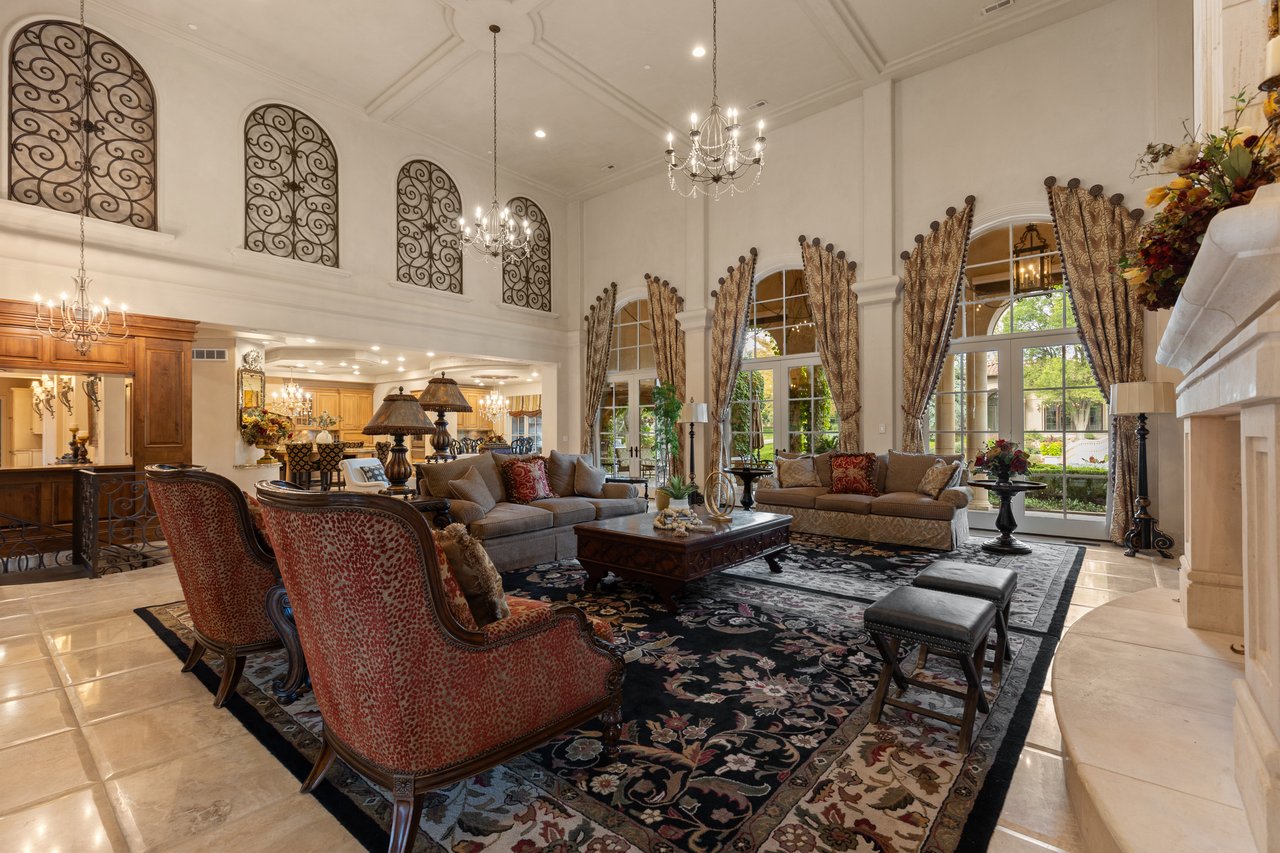 Two-story living room with arched windows, chandeliers, and ornate seating around a floral rug.