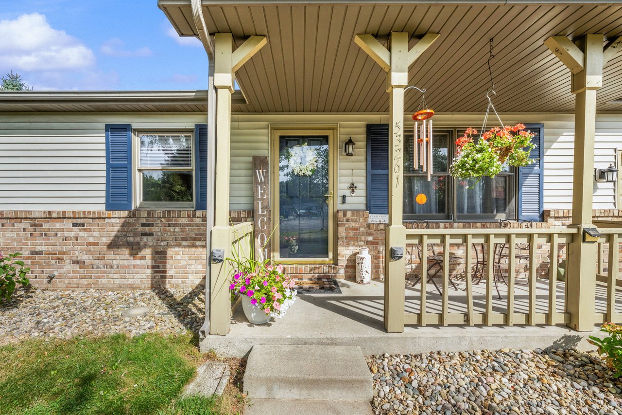 Home entrance with a covered porch, brick façade, hanging flowers, and a welcome sign.
