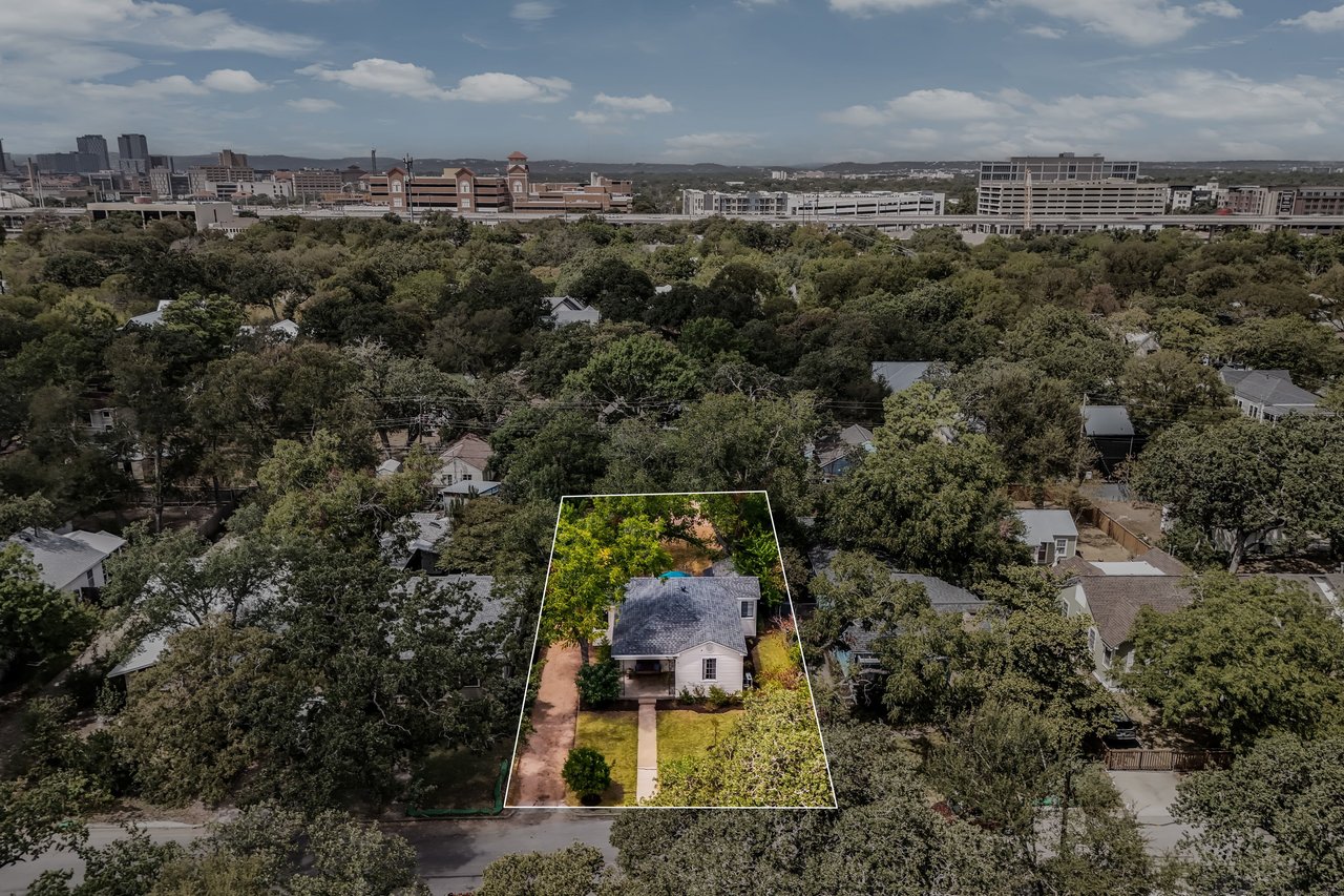 Aerial view of a house and outlined lot among trees, with the city skyline in the distance.