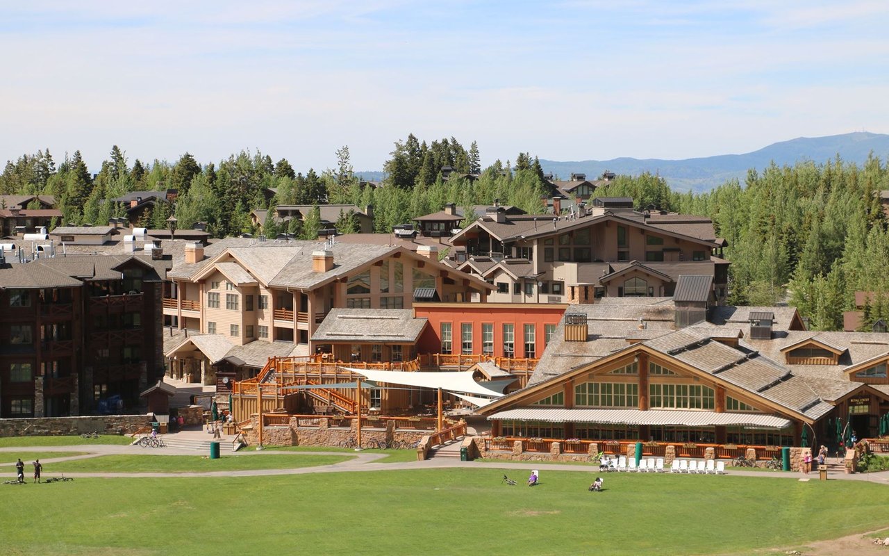 Mountain resort village with timber buildings, a green lawn, evergreen trees, and distant mountains.