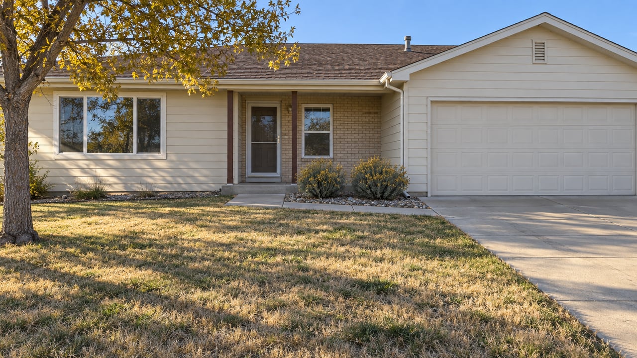 A modest cream-sided ranch home with a buff brick entry, attached garage and a deep, partly dormant front lawn in afternoon sunlight.