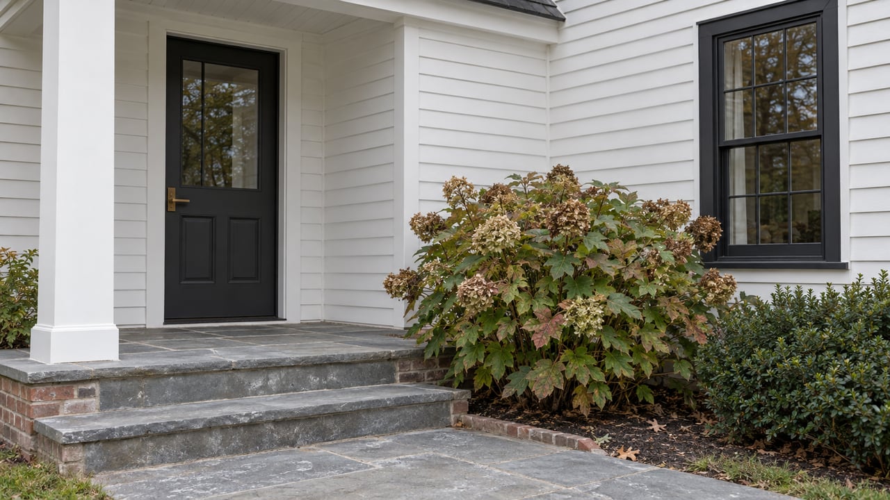 White clapboard house entry with a charcoal paneled door, bluestone steps, and hydrangea beside the path.