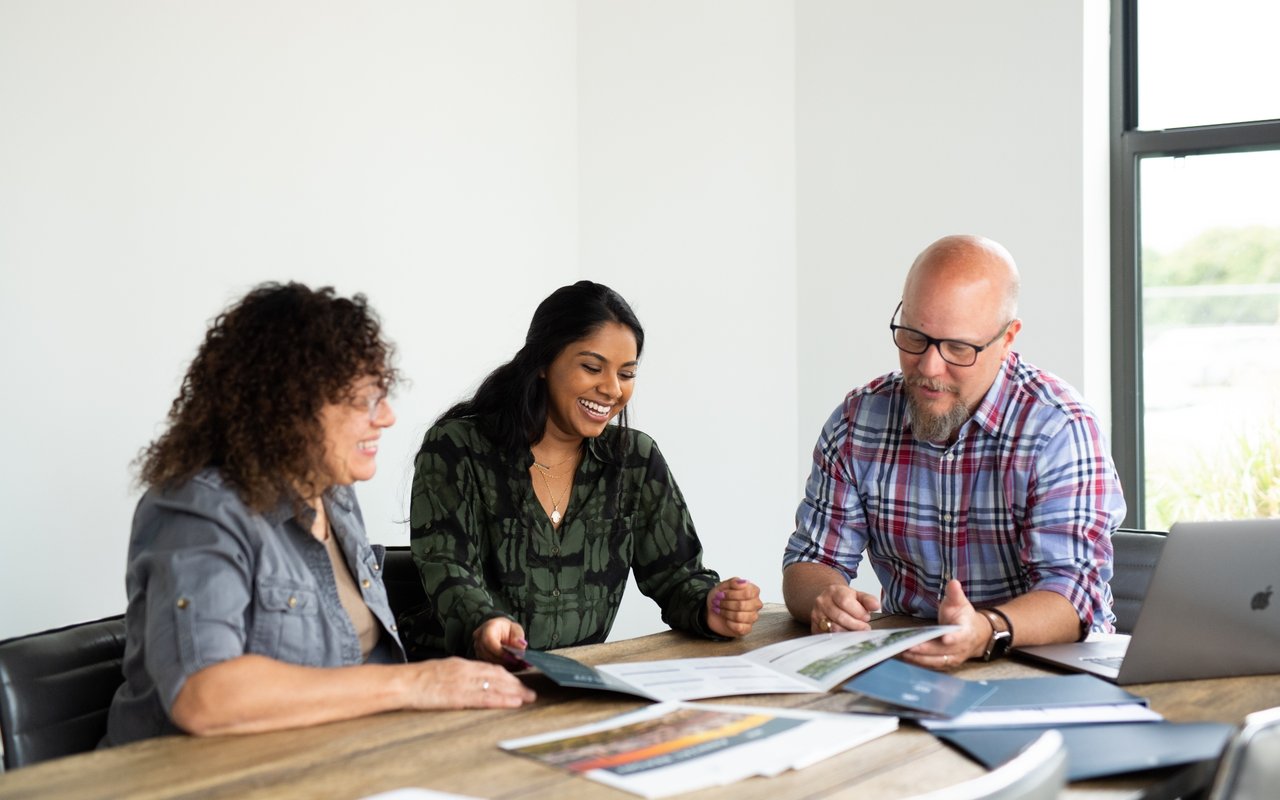Three people review printed materials around a table in a bright office.