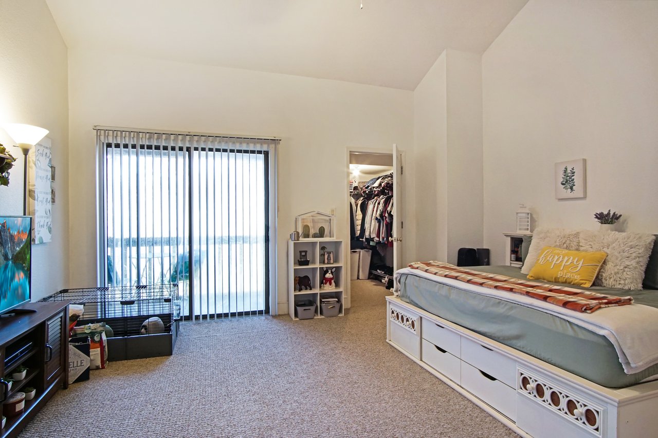 Cozy bedroom with a single bed, fluffy pillows, and a yellow cushion. Open closet full of clothes. Sliding doors with vertical blinds let in natural light. A television, shelves, and pet cage add a personalized touch.