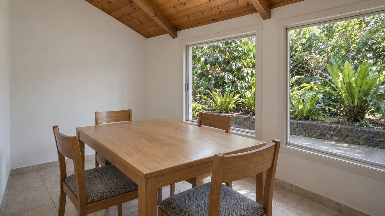 A modest dining corner with a honey-toned wood ceiling, four timber chairs and closed windows overlooking coffee foliage and bird's-nest ferns.