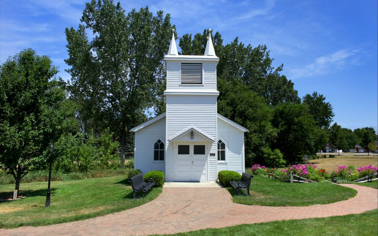 Small white church with twin steeples, surrounded by trees, flower beds, and a curved brick walkway.