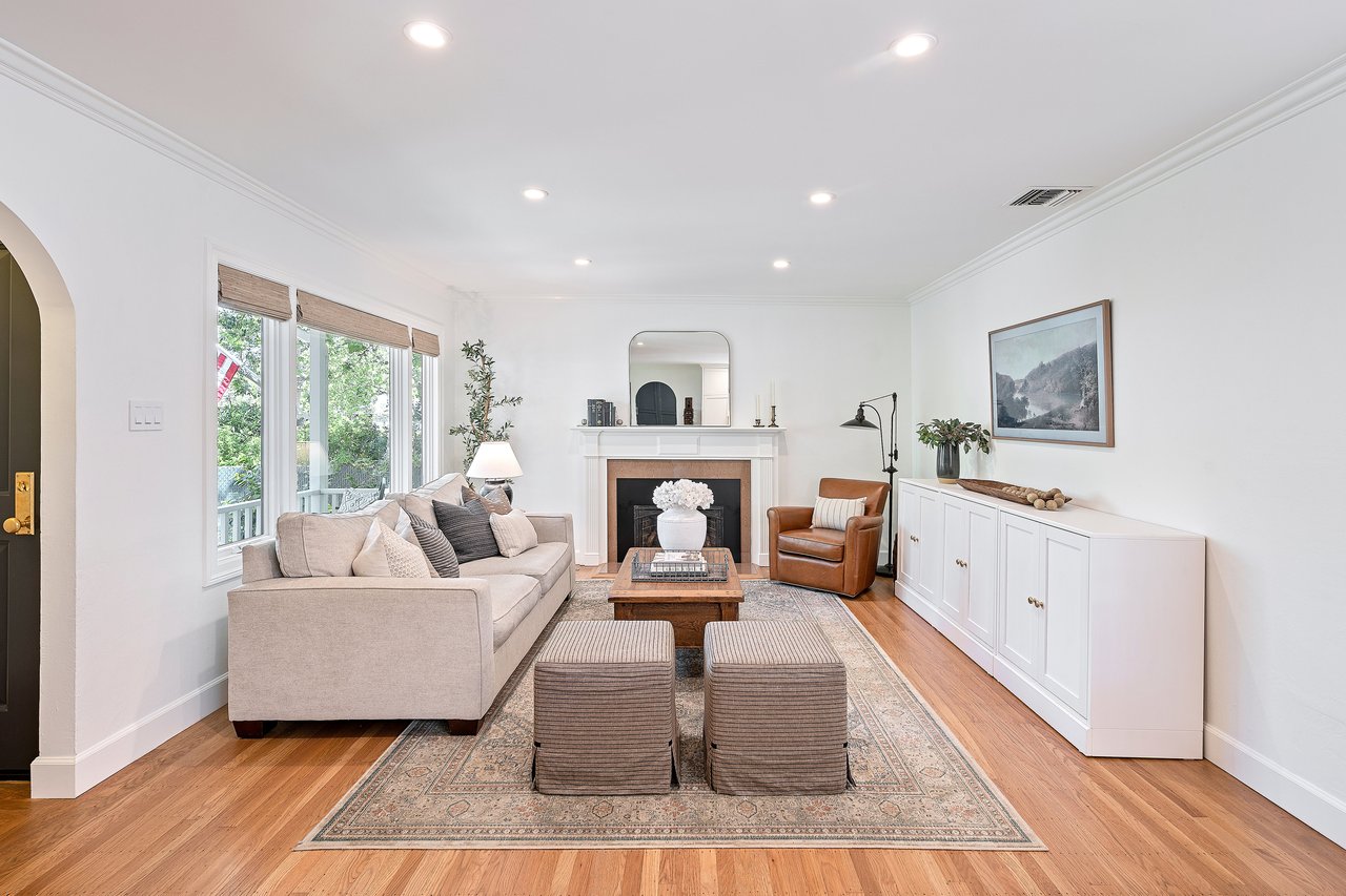 Living room with a fireplace, light sofa, leather armchair, and large windows overlooking greenery.