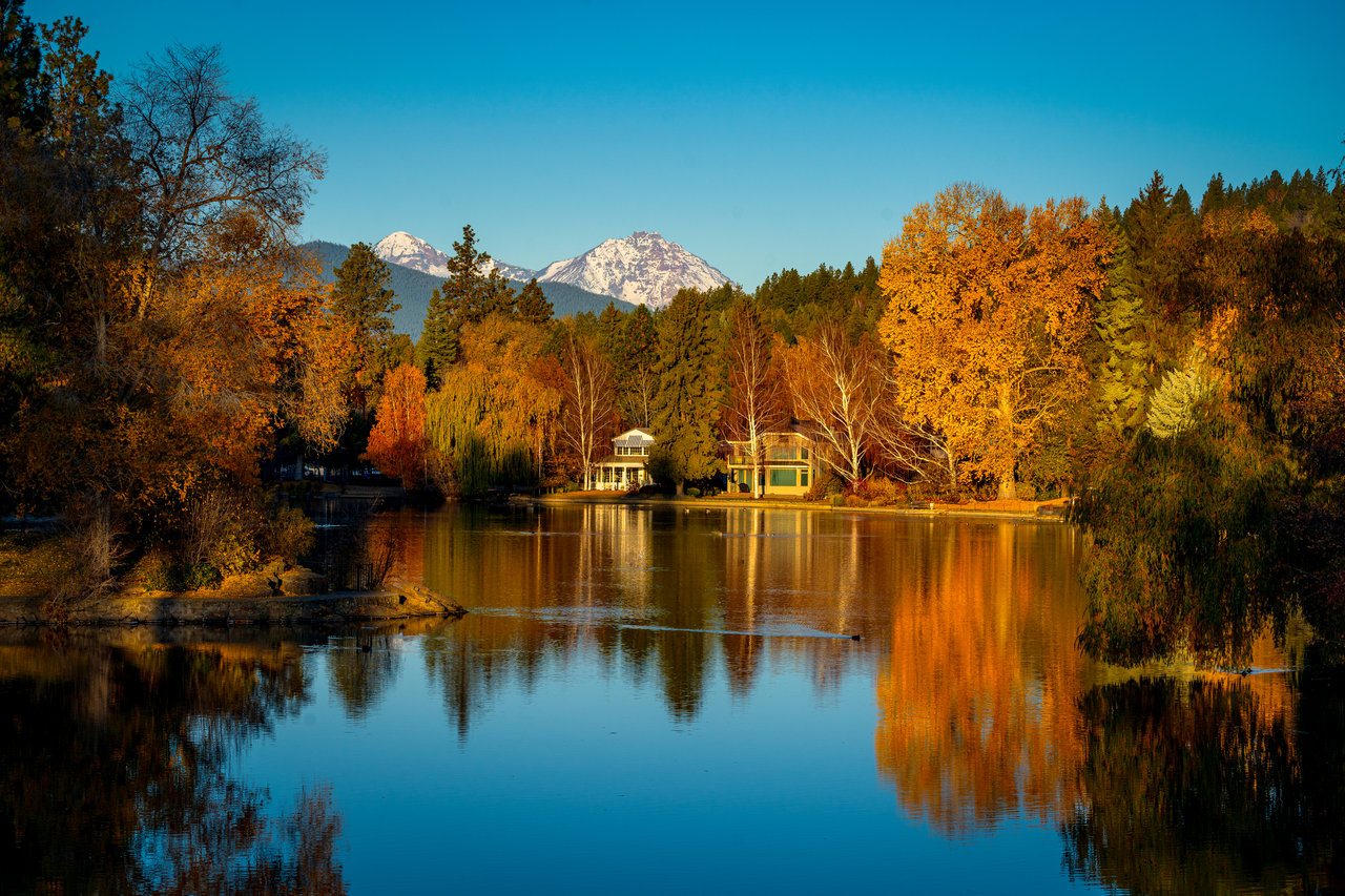 Autumn trees and lakeside homes reflected in a lake, with snow-capped mountains in the distance.