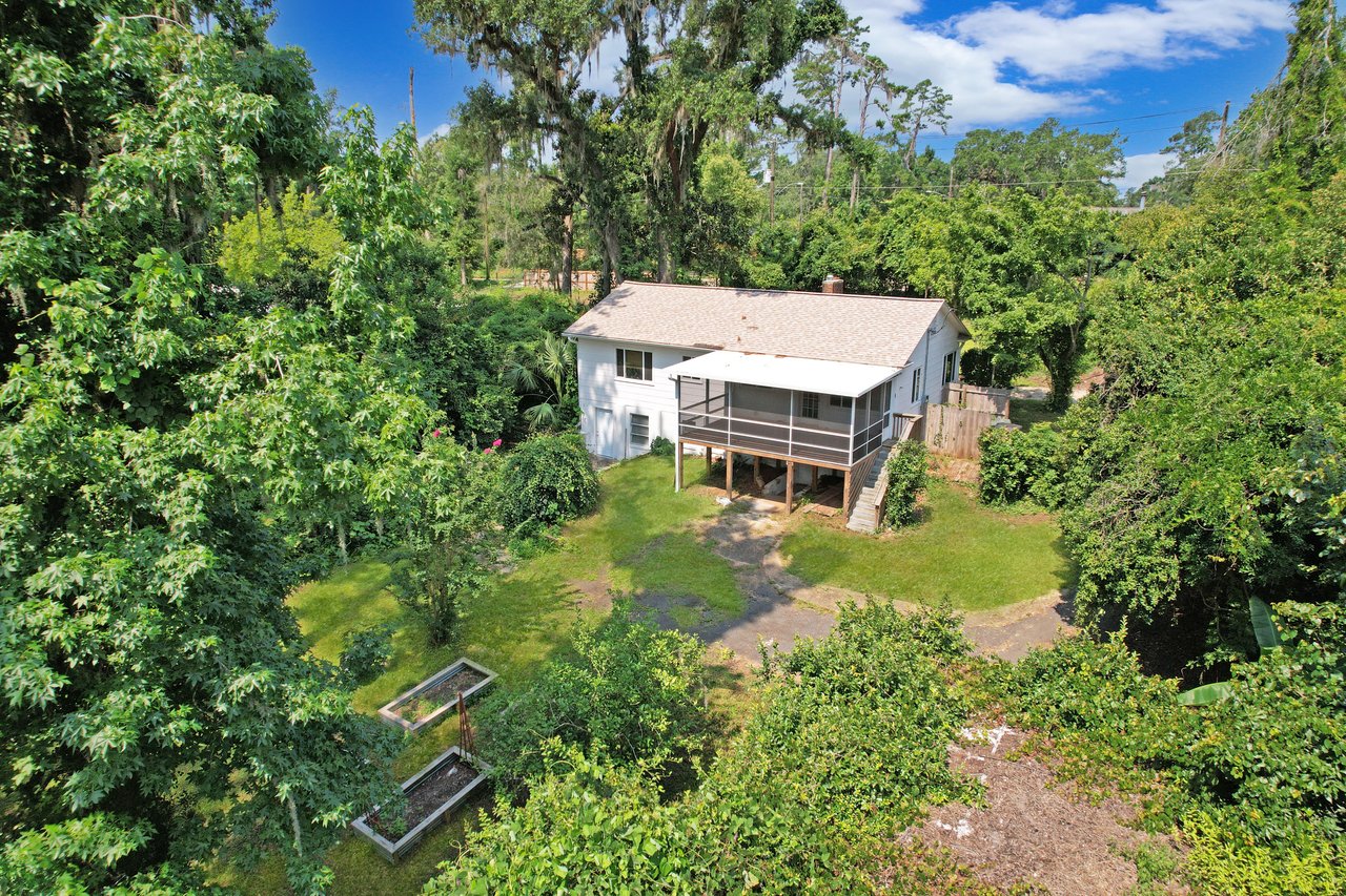 Aerial view of a white house with a screened porch, surrounded by lush green trees and vegetation. Set in a serene, sunlit garden with raised plant beds.