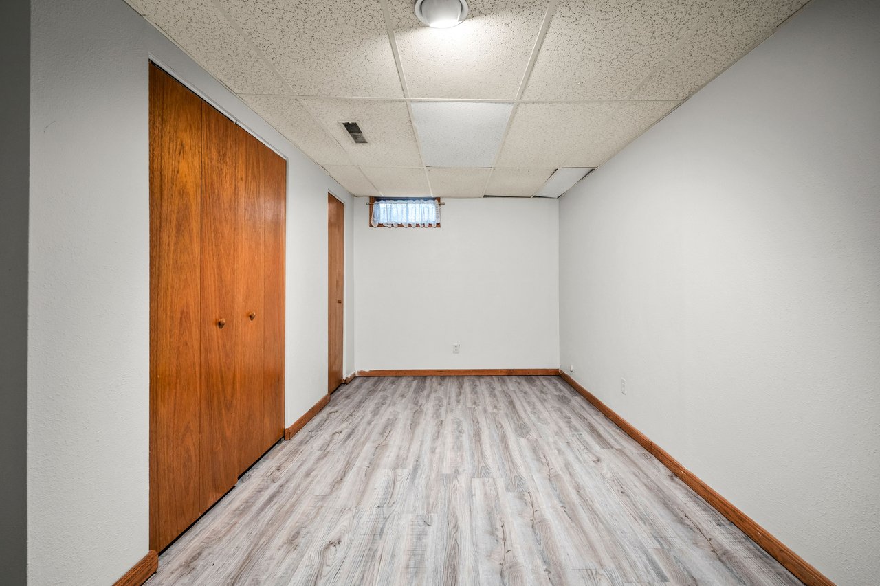 Empty room with gray wood-look flooring, wooden closet doors, and a small curtained window.