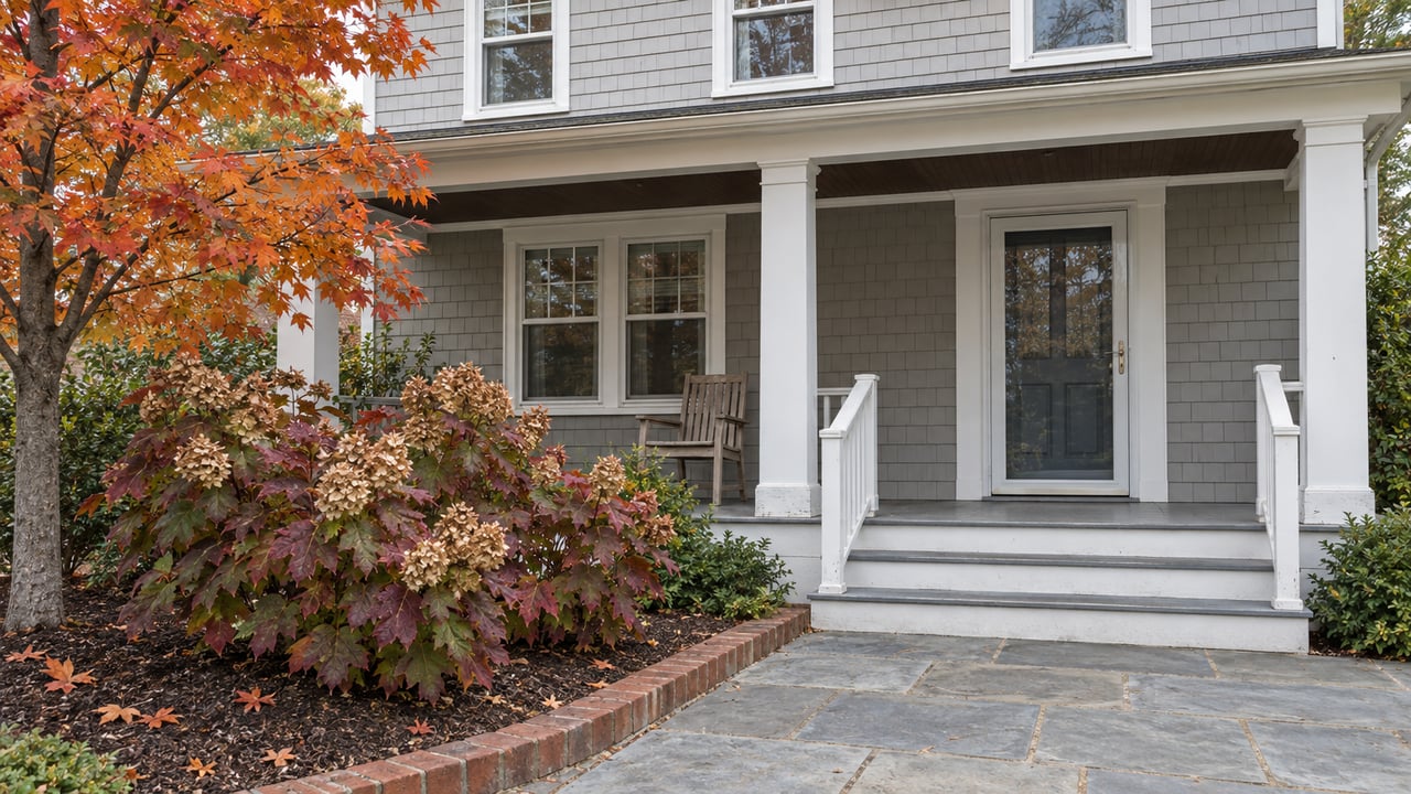 Gray shingle-sided house with a columned porch, stone walkway, and autumn foliage.