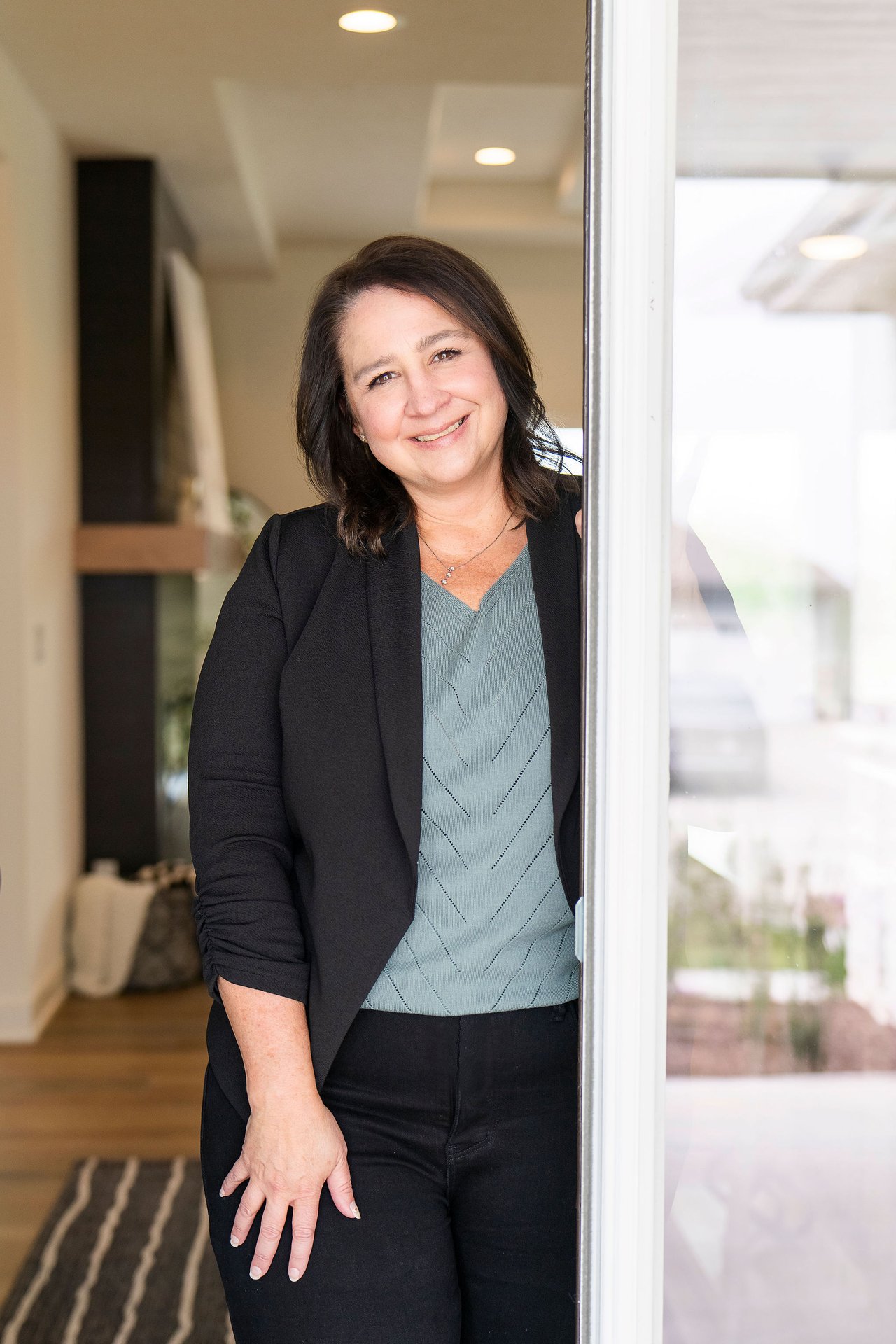 Smiling real estate agent standing beside a glass door in a black blazer and teal top.