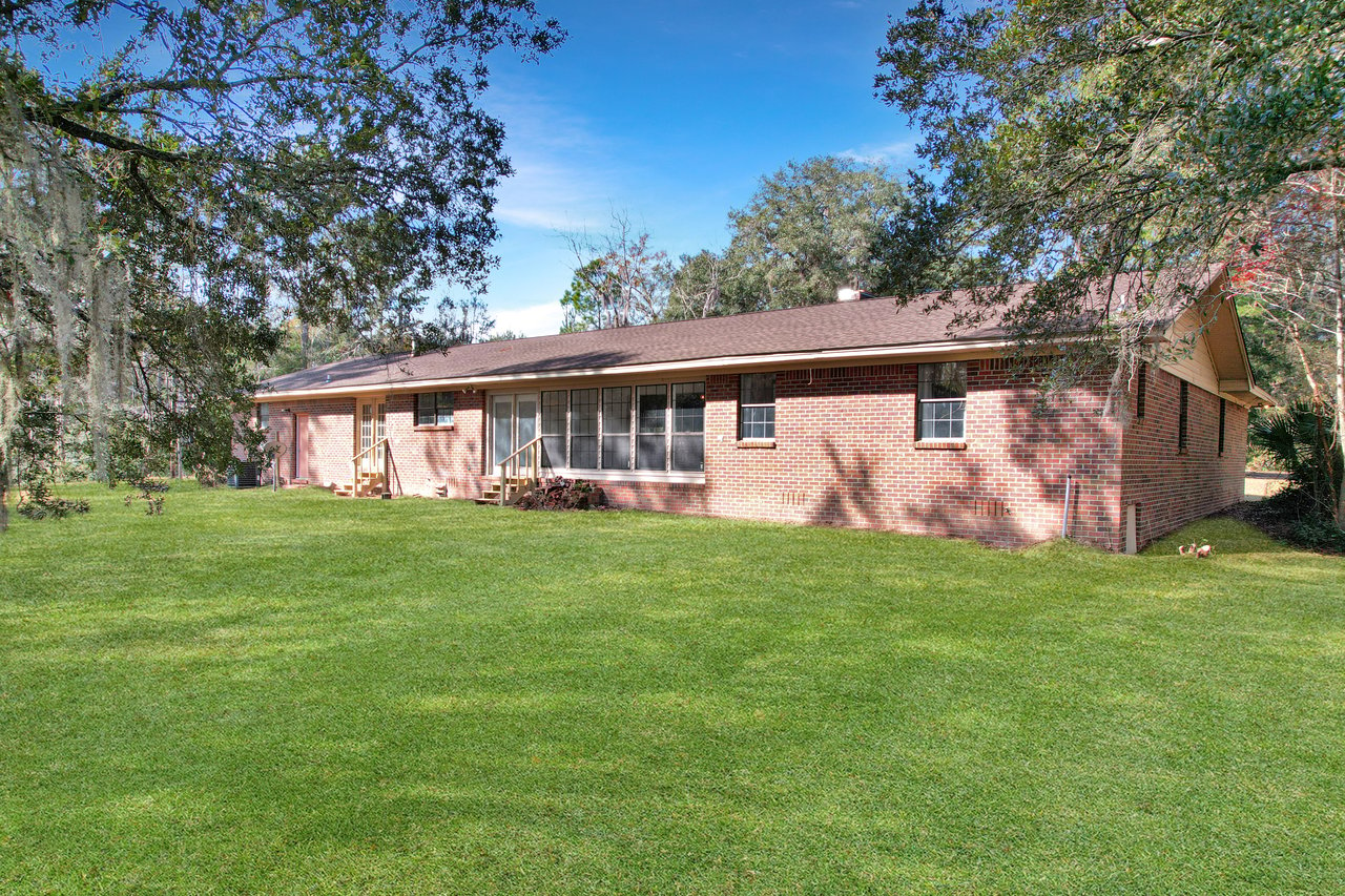 Single-story brick house with a brown roof, large windows, and a green lawn, surrounded by trees under a clear blue sky, conveying a peaceful, suburban feel.