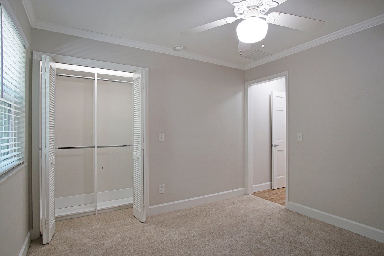 Empty room with beige carpet, white walls, and a ceiling fan. Open closet with white louvered doors and a nearby door leading to another room. Bright and clean.