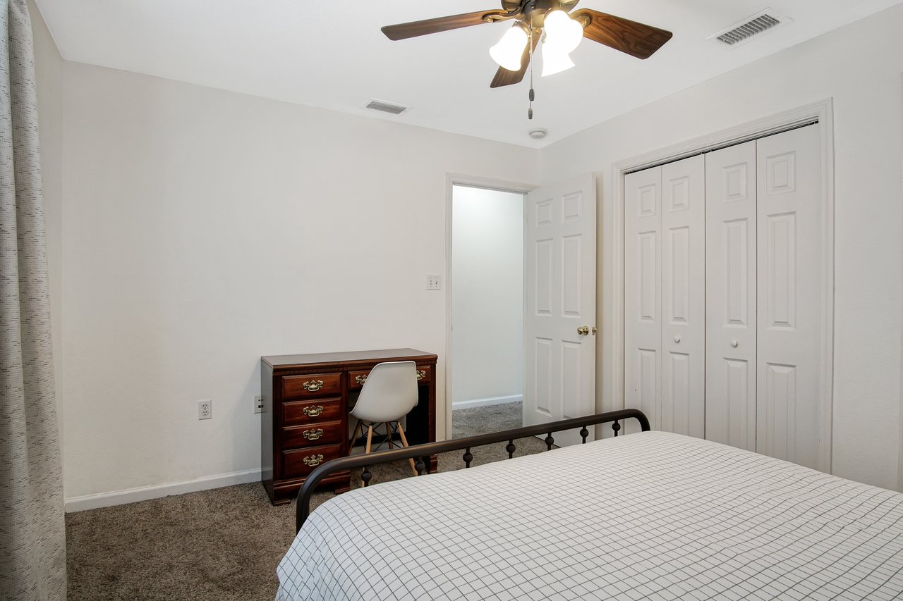 Minimalist bedroom with neutral tones, featuring a grid-patterned bedspread, wooden dresser with a white chair, and a ceiling fan, creating a calm ambiance.