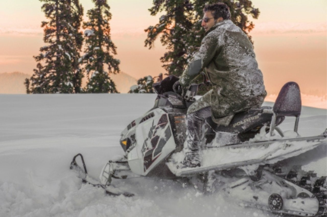 A man riding a Polaris snowmobile through thick, deep powder snow with snow-dusted evergreen trees in the background.