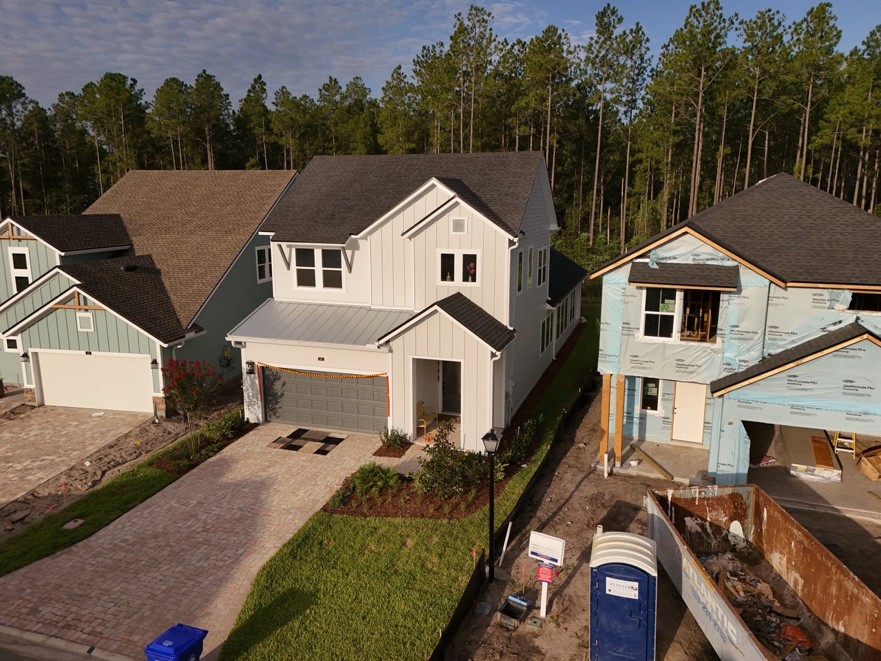 Two-story light-colored home with a brick-paver driveway, beside a neighboring house under construction.