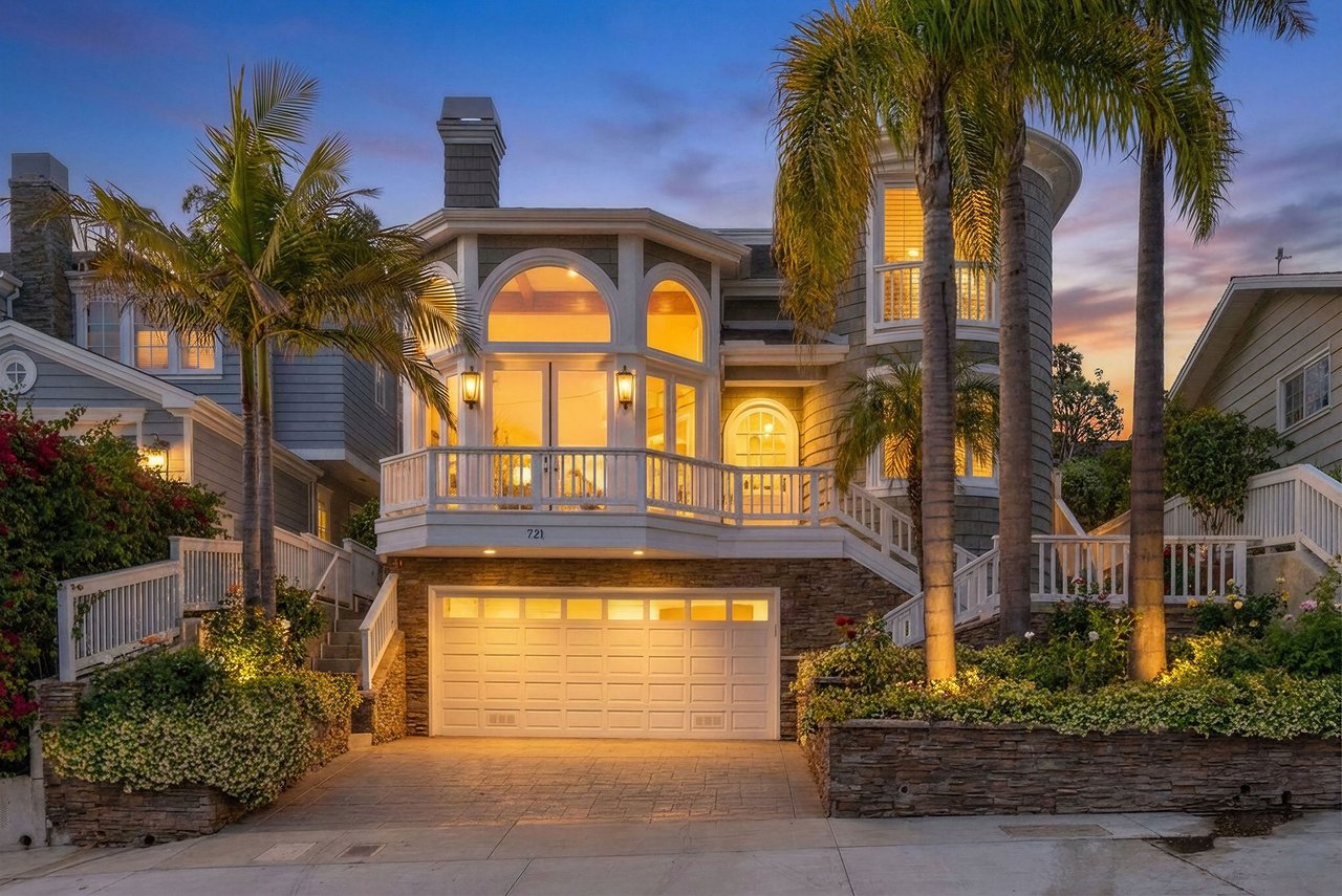 Multi-level gray home with white balconies and palm trees, warmly lit at dusk.