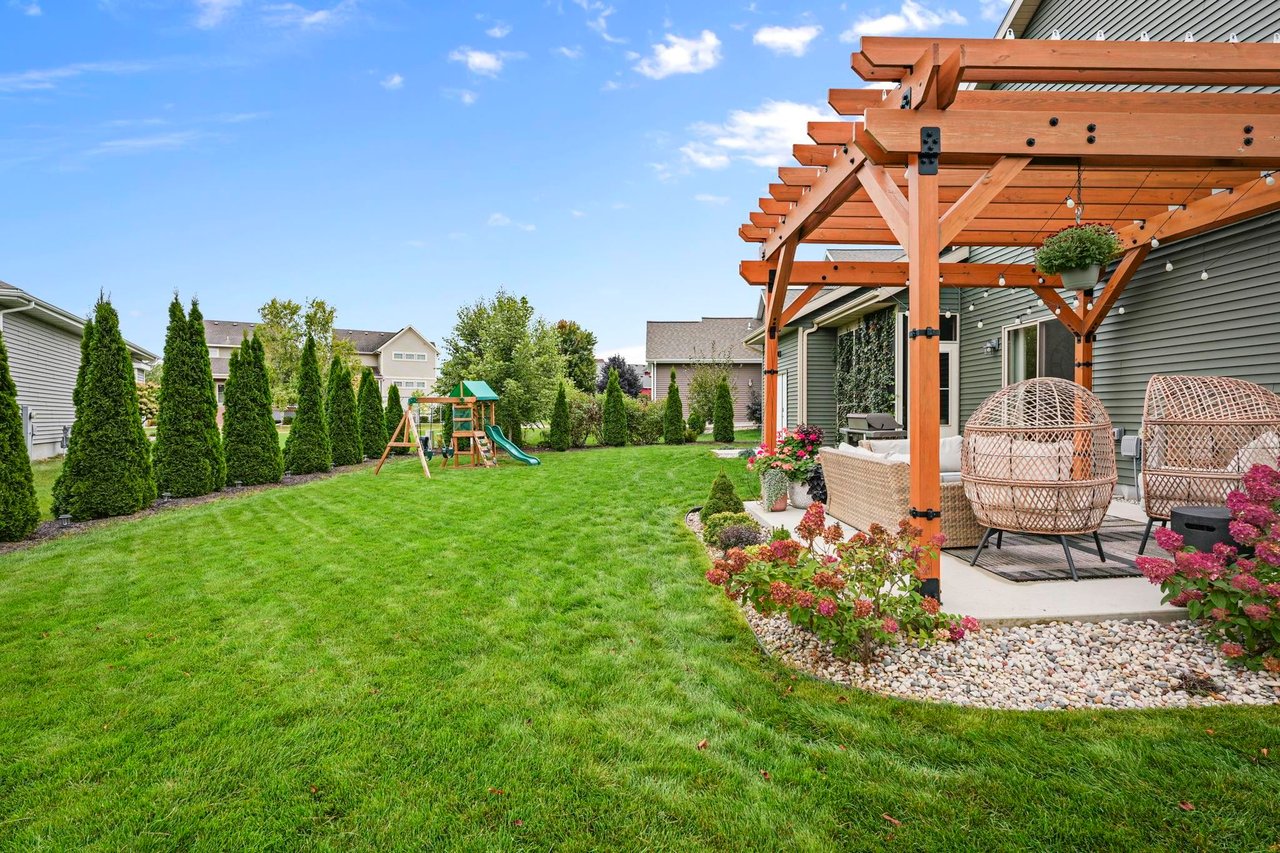A spacious green backyard of a single-family home featuring a wooden pergola over a furnished patio, pink landscaping bushes, a line of privacy trees, and a children's playset in the distance.