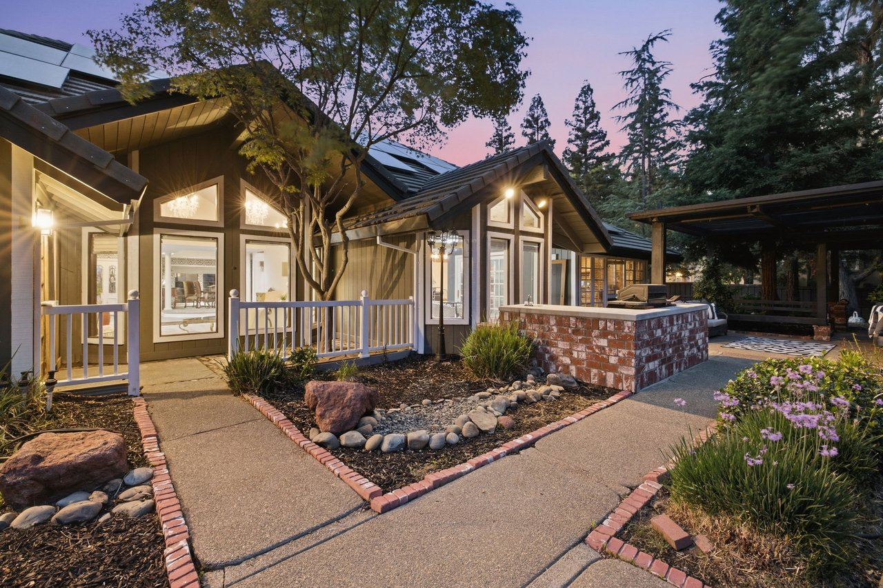 Home at dusk with a brick-edged walkway, landscaped beds, and a covered outdoor kitchen counter.