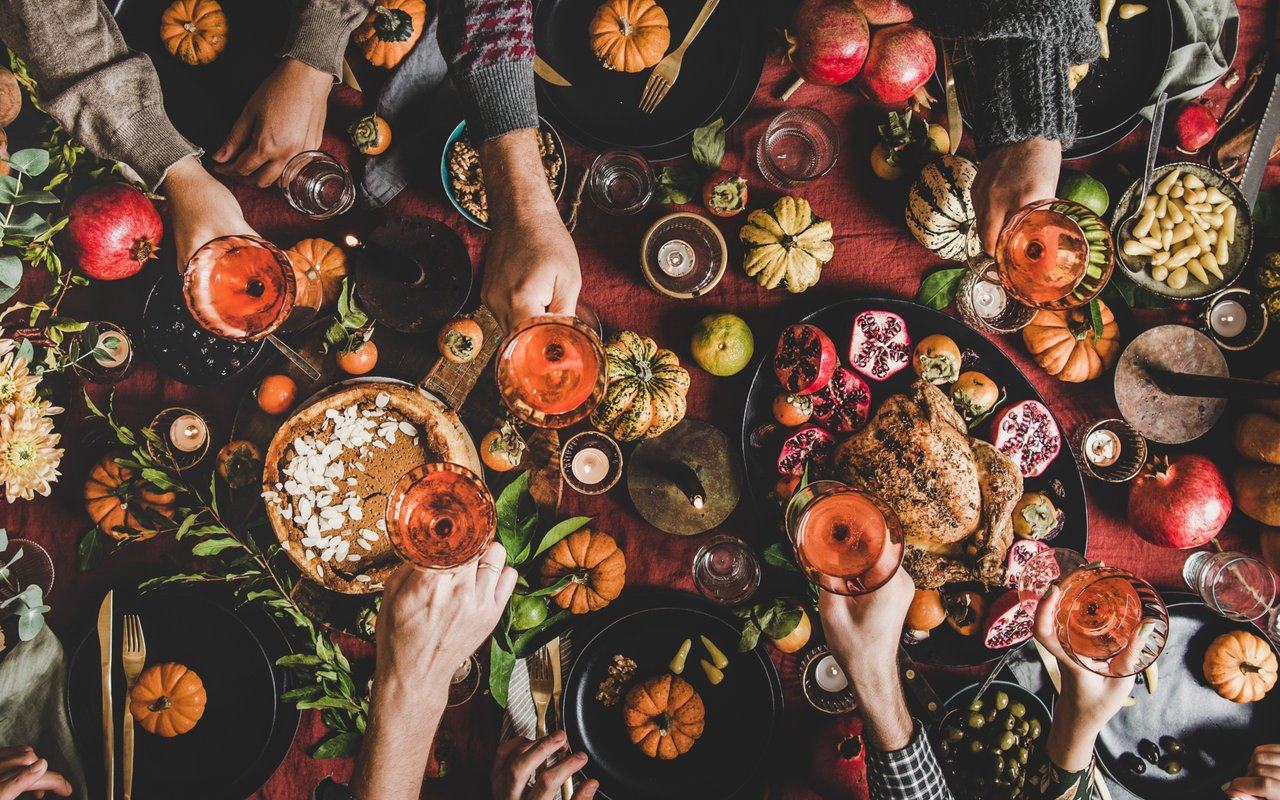 Autumn feast spread with pumpkins, pomegranates, roasted dishes, and hands reaching for wine glasses.