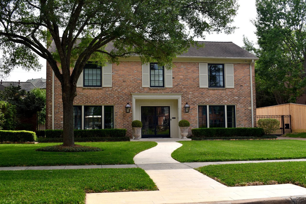 Two-story brick house with a curved stone walkway, broad lawn, and large shade tree.