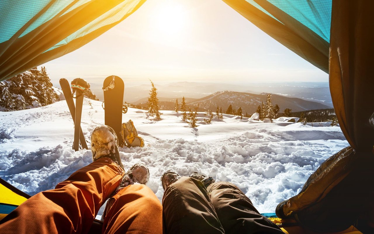 Skiers relax in a tent overlooking snowy mountain ranges at sunset, with skis standing in the snow.