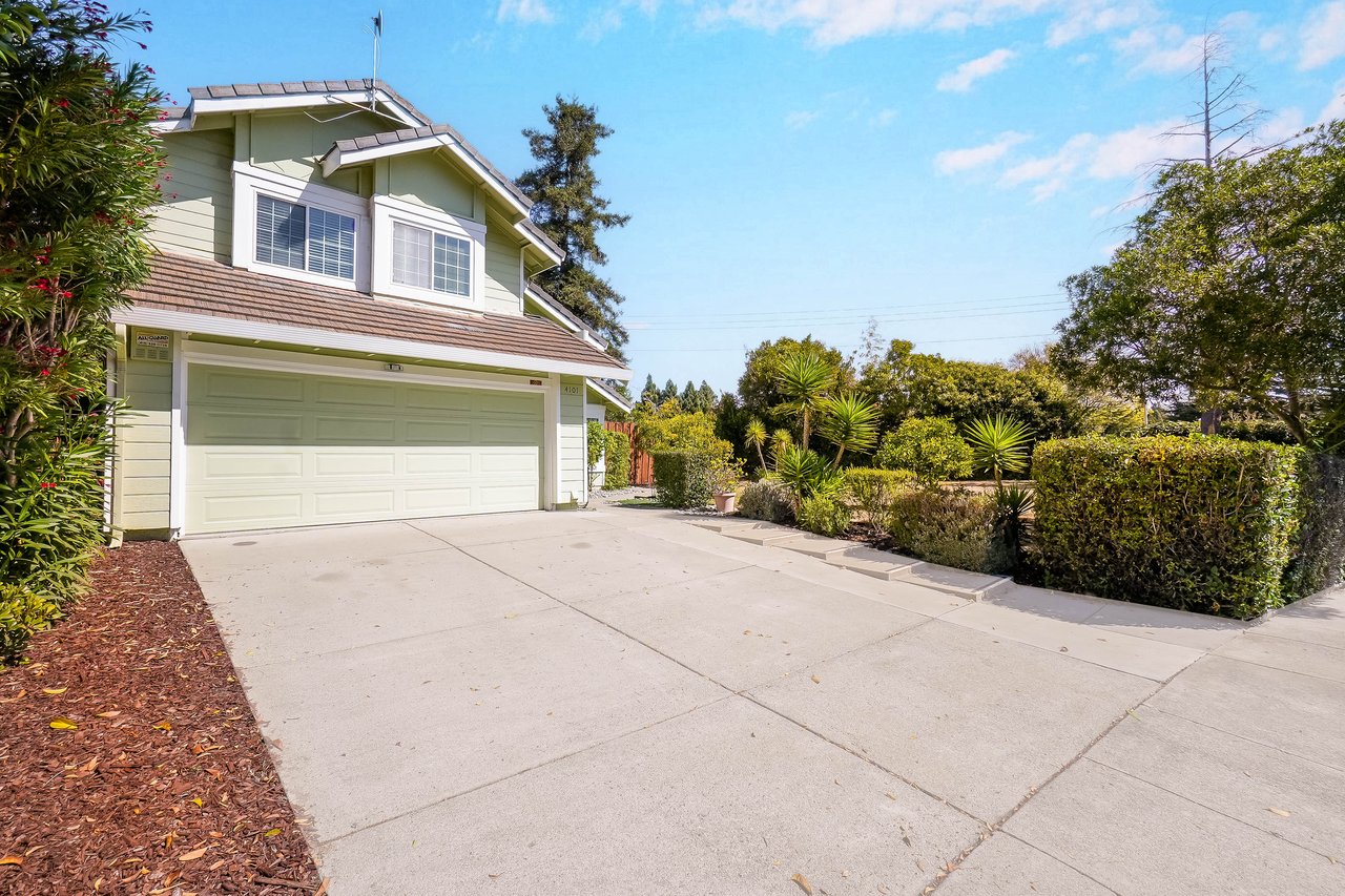 Two-story pale green home with a double garage, broad driveway, and landscaped front yard.