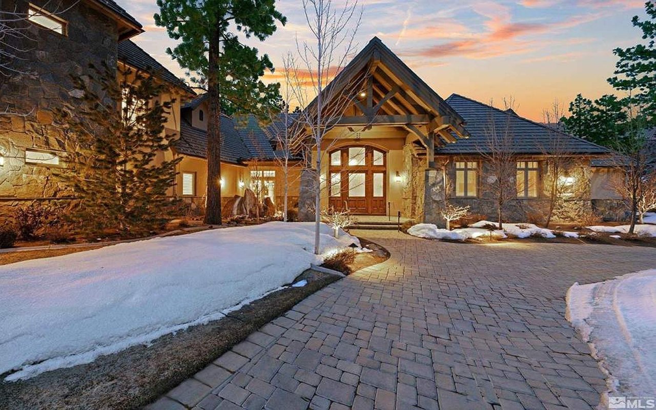 Stone-and-wood home with a snow-covered yard and paved driveway at dusk.