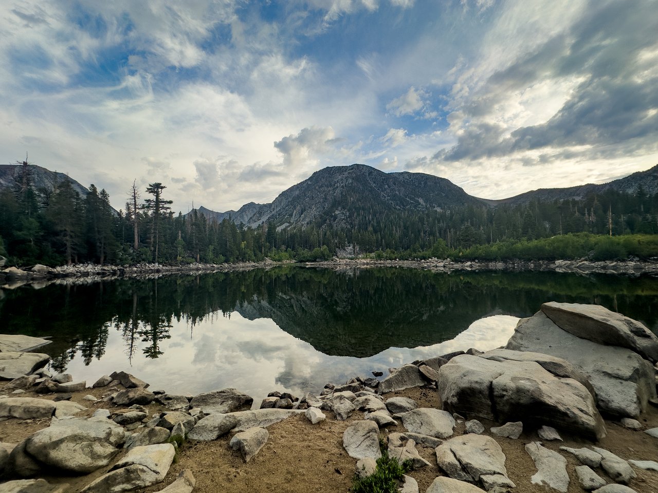 Alpine lake reflecting a forested mountain, with a rocky shore in the foreground.