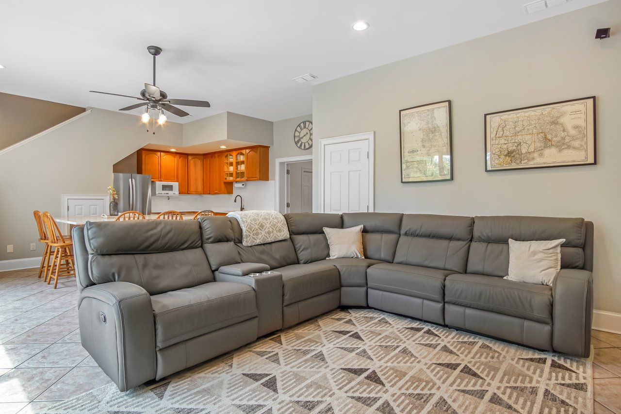 A cozy living room with a large gray sectional sofa, patterned rug, and soft lighting. The kitchen in the background has warm wood cabinets.