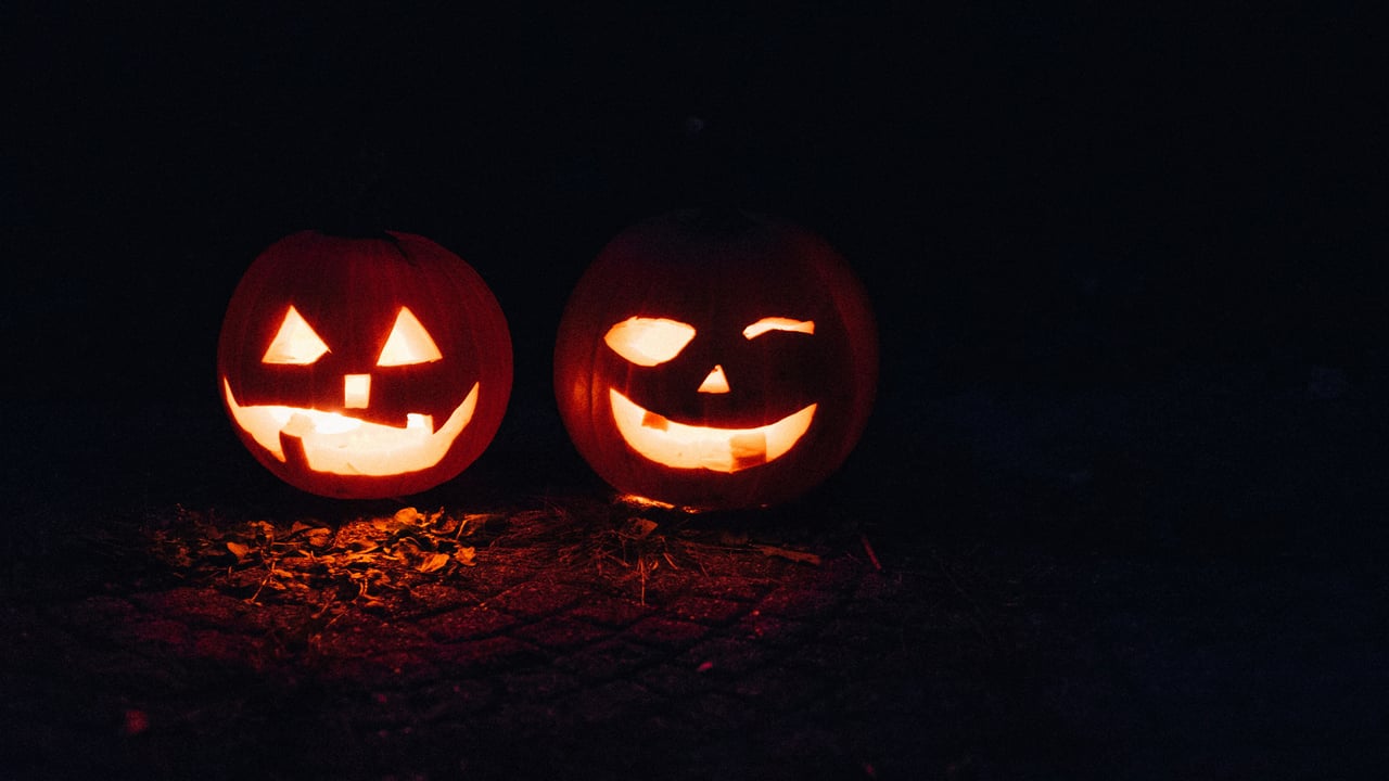 Two glowing jack-o’-lanterns with playful carved faces sit among fallen leaves at night.
