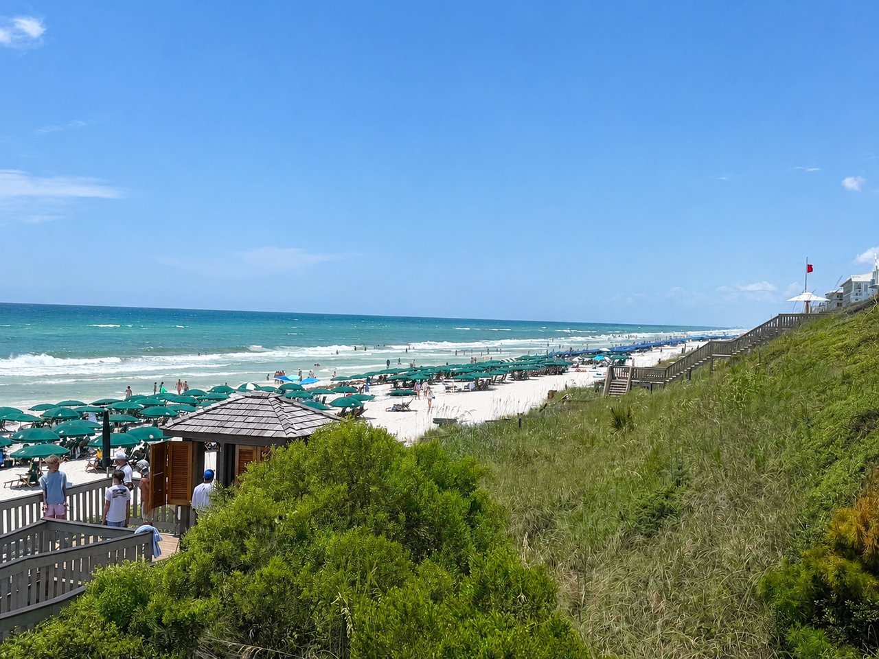 A scenic view from a wooden beach access walkway looking over green dunes and a wooden pavilion, leading down to a white sand beach dotted with green umbrellas and the ocean under a bright blue sky.