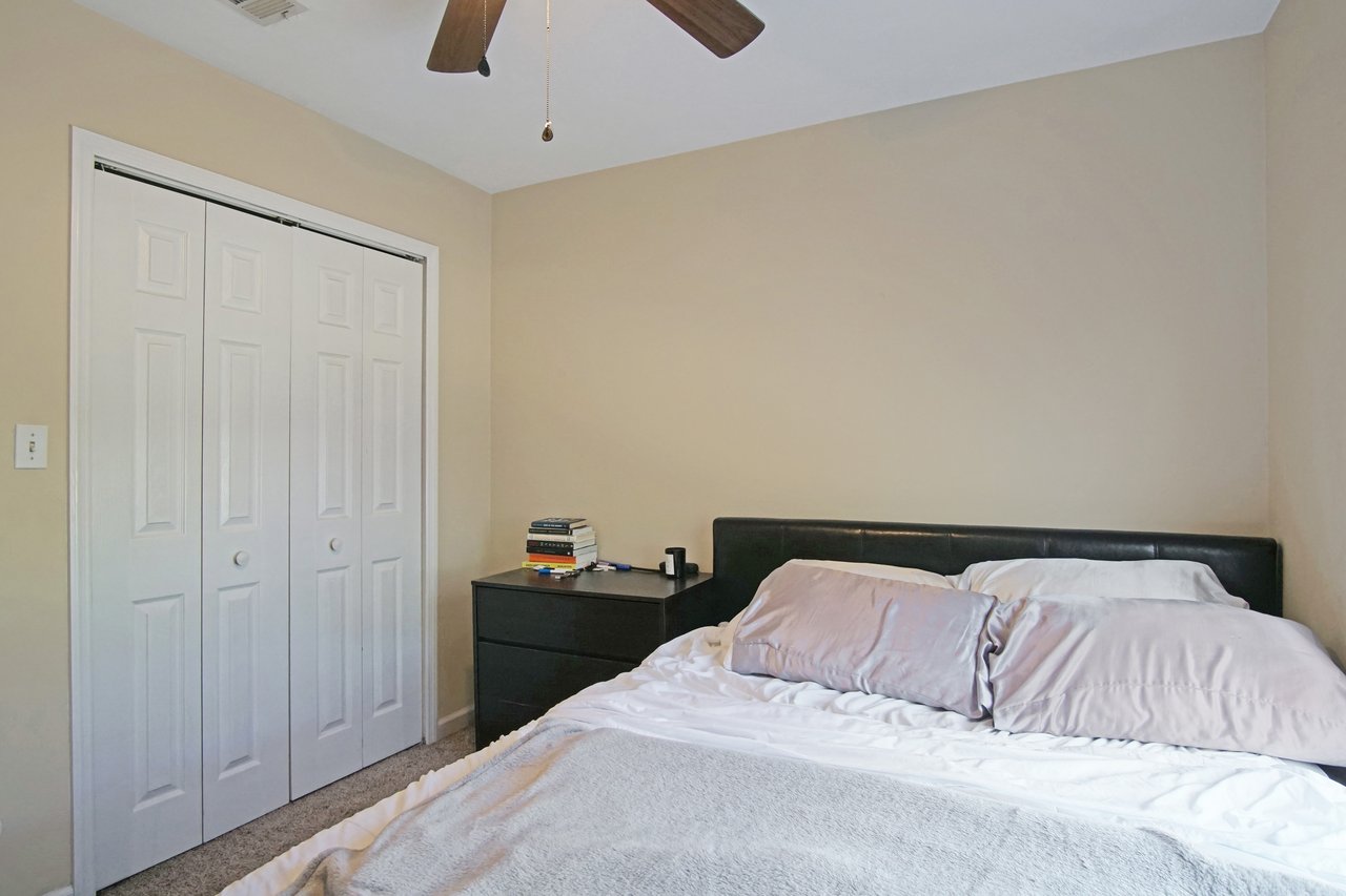 Bedroom at 529 Collinsford Road, Tallahassee, Florida, featuring beige walls, carpet flooring, and a ceiling fan. The room includes a bed with light bedding, a black dresser with books on top, and a white double-door closet for storage.