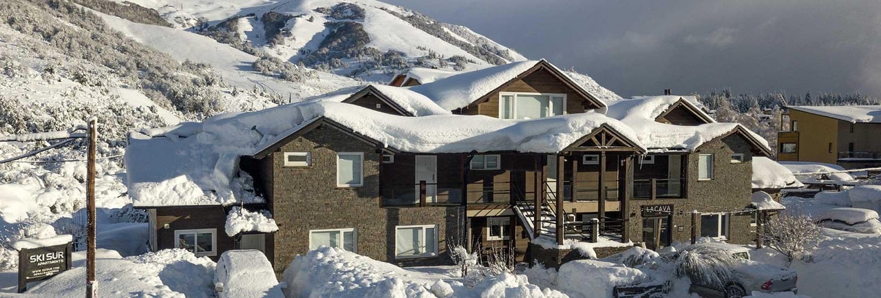 Stone-and-timber lodge beneath snowy mountain slopes, with deep snow covering its roof.