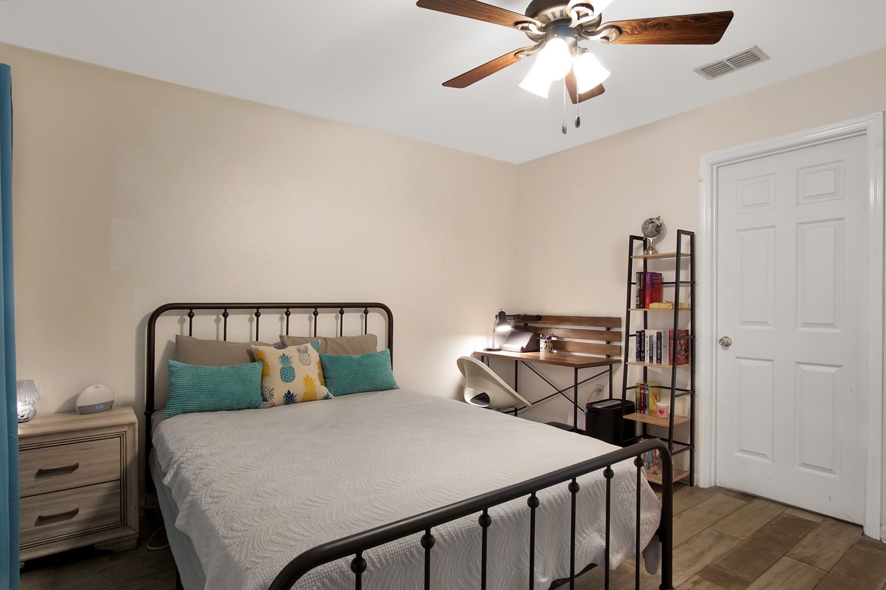 Cozy bedroom with cream walls and wood flooring. Features a metal-framed bed with colorful pillows, a small nightstand, and a desk with a lamp. A ceiling fan hangs above.