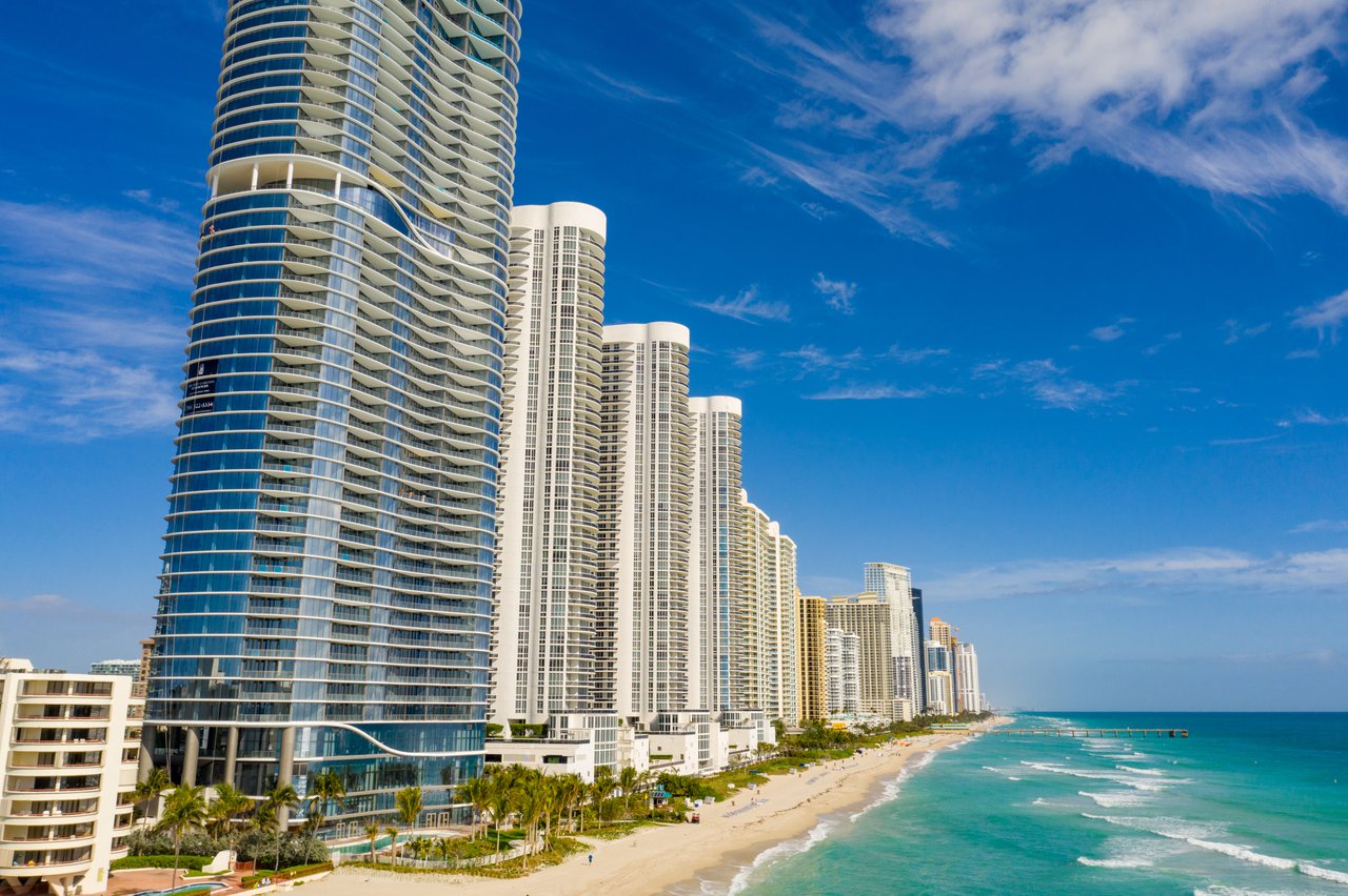 Beachfront condominium towers line a sandy shore beside turquoise ocean water.