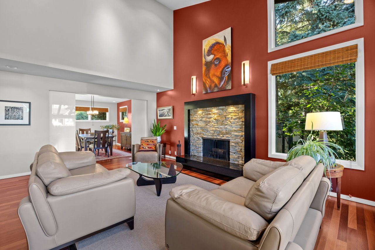 Living room with a stone fireplace, vaulted ceiling, red accent wall and large windows overlooking trees.