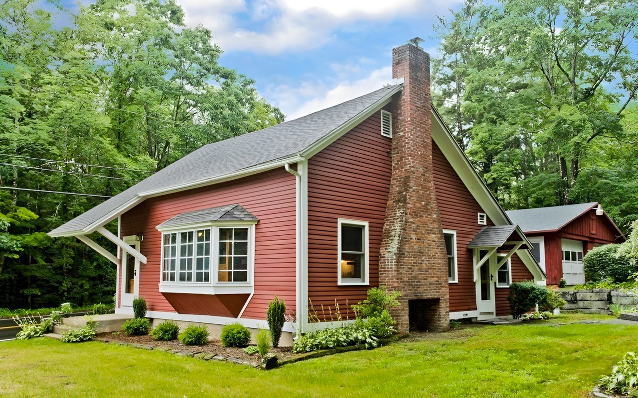 Red single-family home with white trim, a large brick chimney, and a bay window among leafy trees.