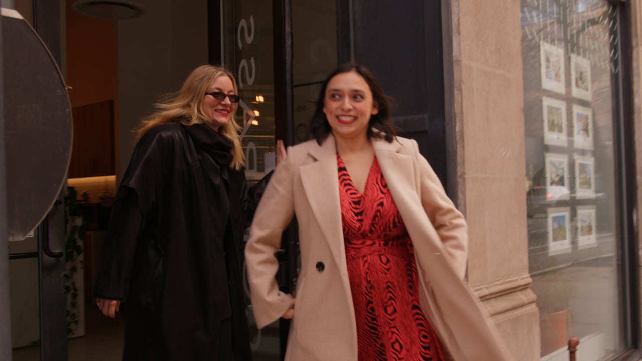 Two women outside a storefront, one in a red patterned dress and pale coat.