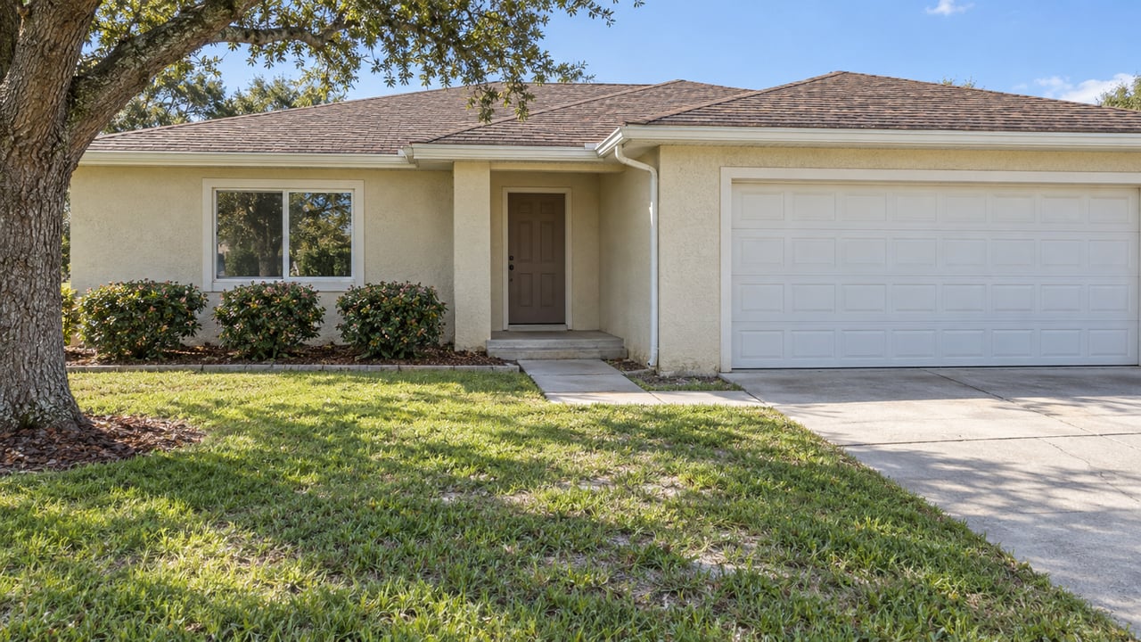 Single-story ivory stucco home with brown shingle roof, white garage door, concrete approach and sunlit lawn.