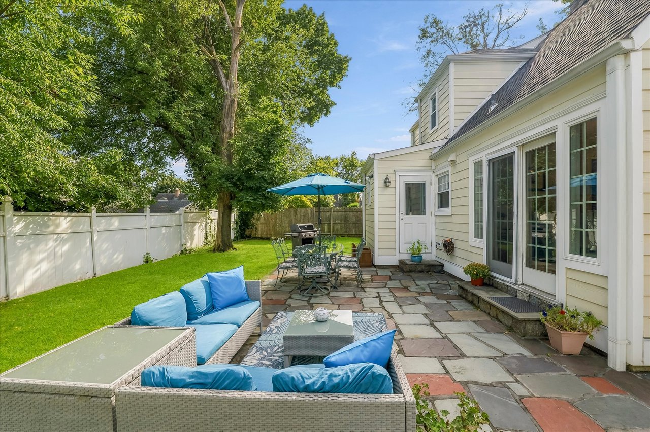Backyard patio with blue-cushioned seating, stone paving, dining set, and a large shade tree.