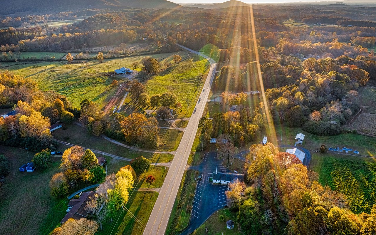A rural road winds past homes and fields among autumn-colored trees and rolling hills.