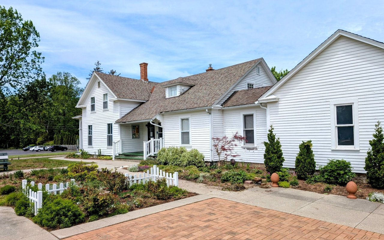 White multi-wing house with a landscaped garden, picket fence, and brick-paved driveway.